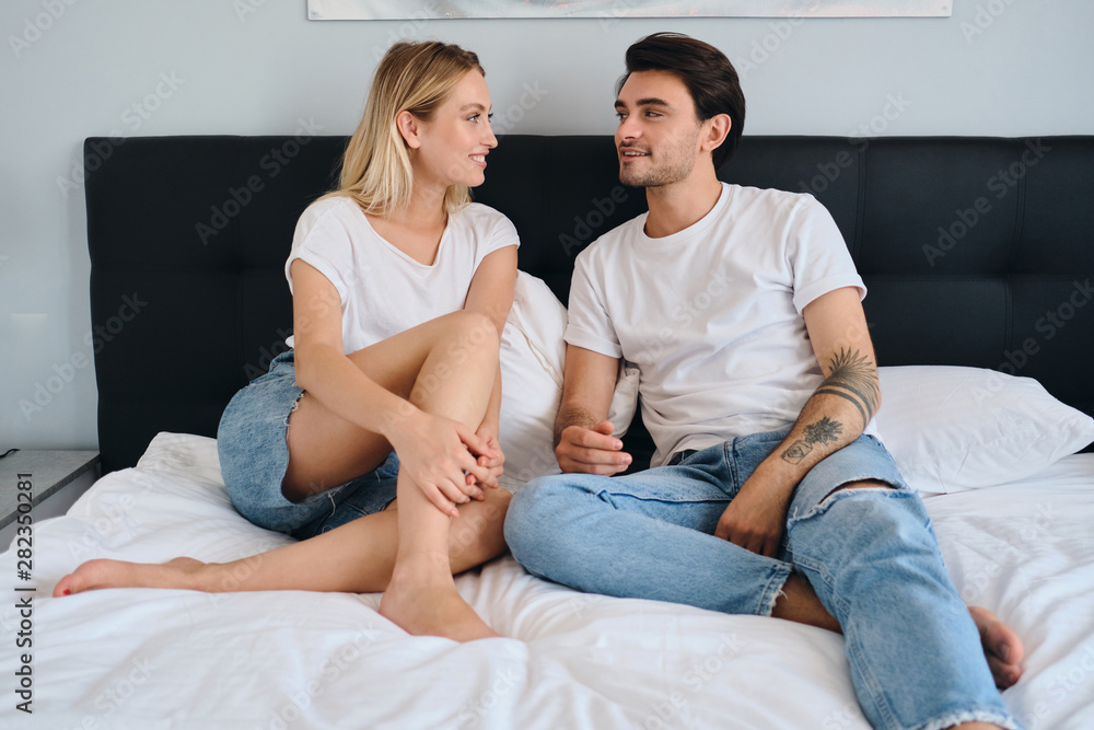 Young attractive brunette man and beautiful smiling blond woman in white T-shirts talking and dreamily looking at each other on bed in modern hotel Young attractive brunette man and beautiful smiling blond woman in white T-shirts talking and dreamily looking at each other on bed in modern hotel