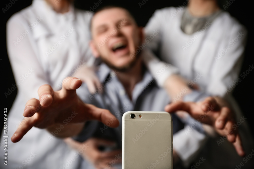 Emotional man reaching for smartphone while medical workers holding him back on black background, closeup. Addiction concept - obrazy, fototapety, plakaty 