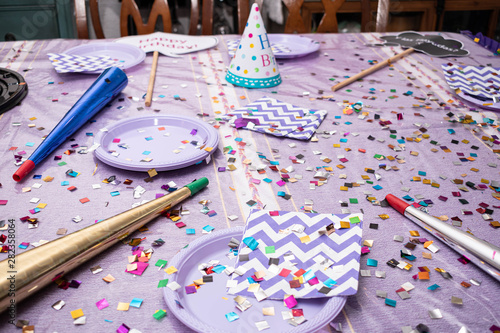 Table with elements for a party like birthday hat, confetti, trumpets, disposable plates and napkins on a purple tablecloth