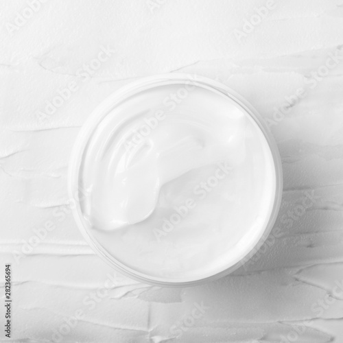 Open white jar with white face cream on a background of smeared cream. Cosmetic moisturizing product. Top view, flat lay.