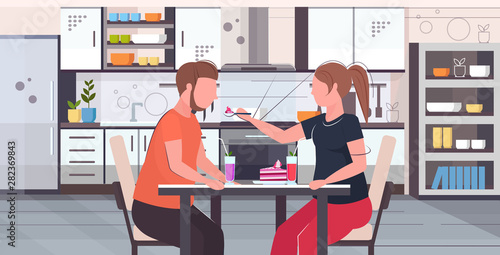 obese couple sitting at table eating sweet tasty cake overweight woman feeding fat man with spoon unhealthy nutrition obesity concept modern kitchen interior flat portrait horizontal