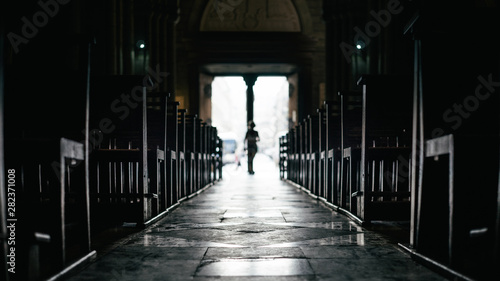 Rows of bench inside a silent church with silhouette of passerby moving in from exit, in France, Europe