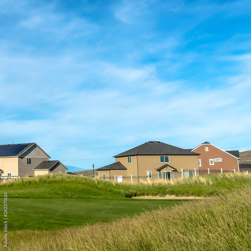Square frame Grassy terrain and houses under blue sky with white clouds on a sunny day Square frame Grassy terrain and houses under blue sky with white clouds on a sunny day