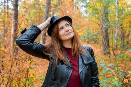 Beautiful red-haired girl in a red dress, black leather jacket, hat looking into the distance in the autumn forest. Girl in the autumn forest. Look away. Horizontal photography