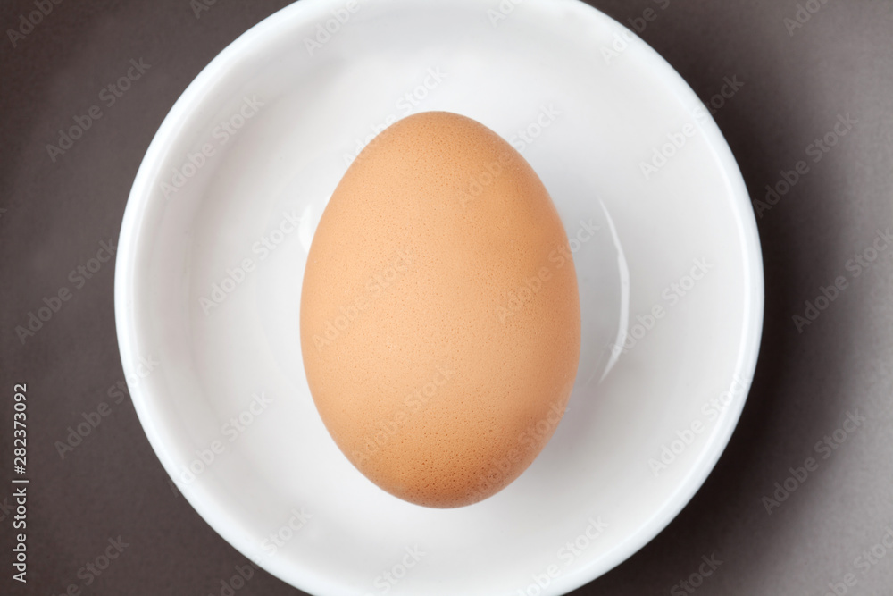 One raw egg on white plate on brown background from top view with space for copy.