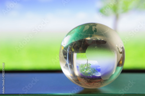 World globe crystal glass reflect in green wide grassland, tree and blue sky with clouds gloss on table beside the window. Global business and economy. Environmental conservation or ecology.