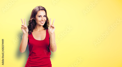 Portrait of young happy smiling beautiful woman in casual clothing, showing two fingers or victory gesture, over yellow color wall background. Happy girl in red dress.