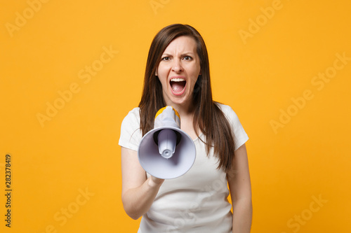 Portrait of angry young woman in white casual clothes looking camera and scream in megaphone isolated on bright yellow orange wall background in studio. People lifestyle concept. Mock up copy space.