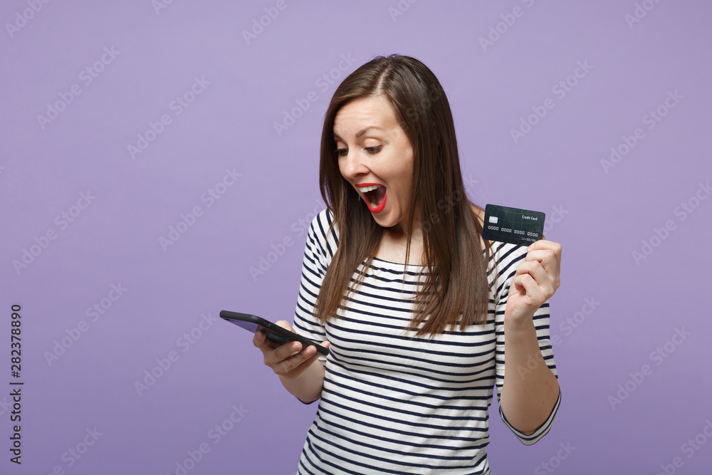 Young brunette woman girl in casual striped clothes posing isolated on violet purple background studio portrait. People lifestyle concept. Mock up copy space. Hold in hand credit bank card cellphone.
