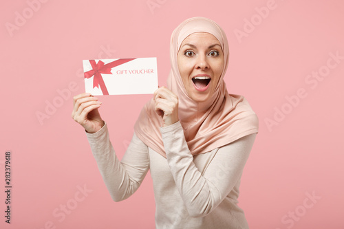 Shocked amazed young arabian muslim woman in hijab light clothes posing isolated on pink background. People religious Islam lifestyle concept. Mock up copy space. Holding in hands gift certificate.