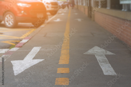 Wallpaper Mural Two white arrow sign and yellow line shown on surface cement footpath and blurred group car parking on sideroad. Blurry people walking on walkway with symbol direction and brown brick wall background. Torontodigital.ca