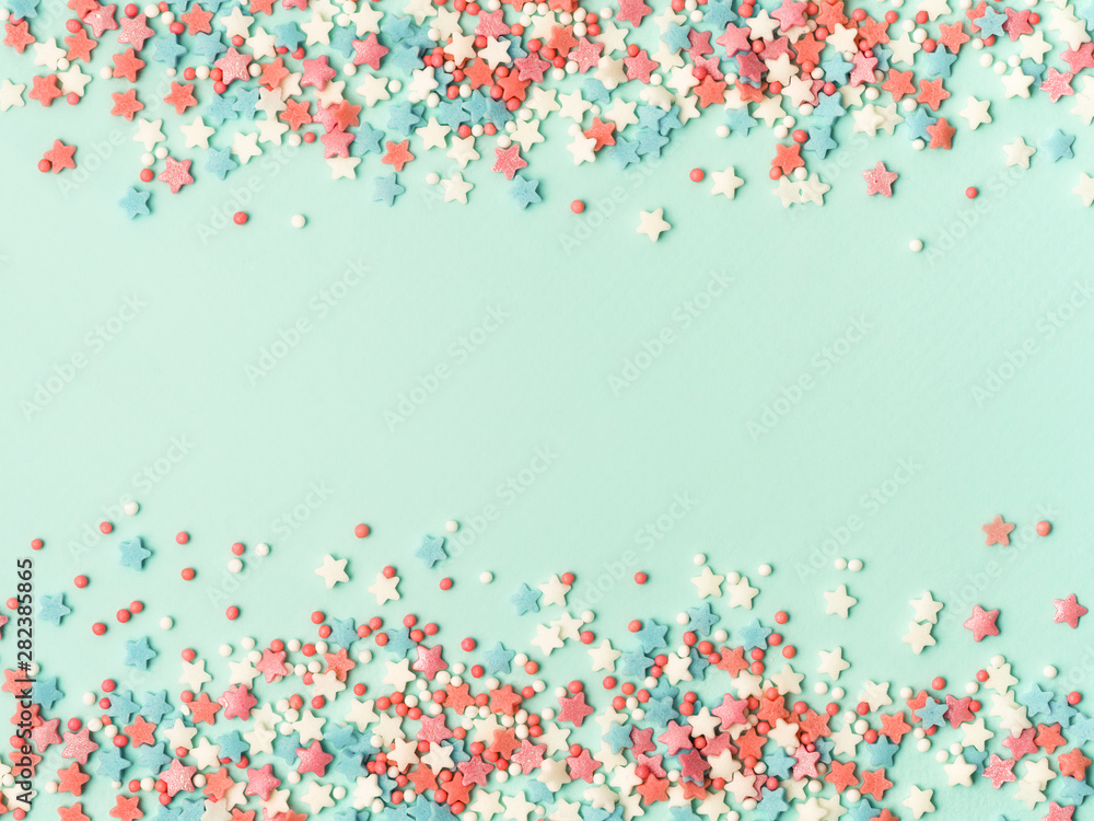Festive border frame of colorful pastel sprinkles on blue background with copy space in center. Sugar sprinkle dots and stars, decoration for cake and bakery. Top view or flat lay. - obrazy, fototapety, plakaty 