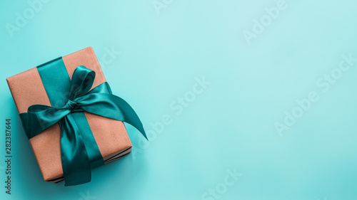 Gift box in craft wrapping paper and green satin ribbon on turquoise blue background, copy space right. Beautiful Christmas, New Year or Birthday present, flat lay or top view. Banner photo