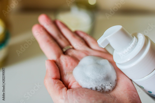 Face cleanser on female hand and cosmetics foam pump container. White blurred background.