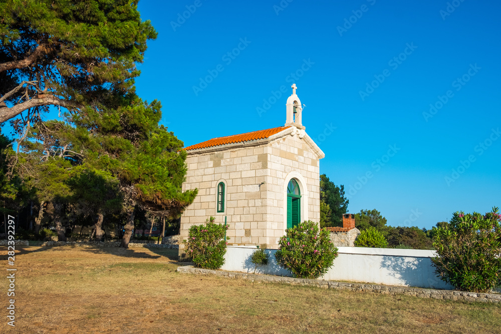 Croatia, island of Dugi Otok, beautiful small church of Veli Rat on the stone shore among the pines and flowers on sunny day, Mediterranean landscape
