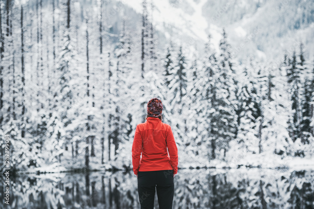 One girl hiker stands in front of a winter lake looking to the snowy landscape. Rear view.