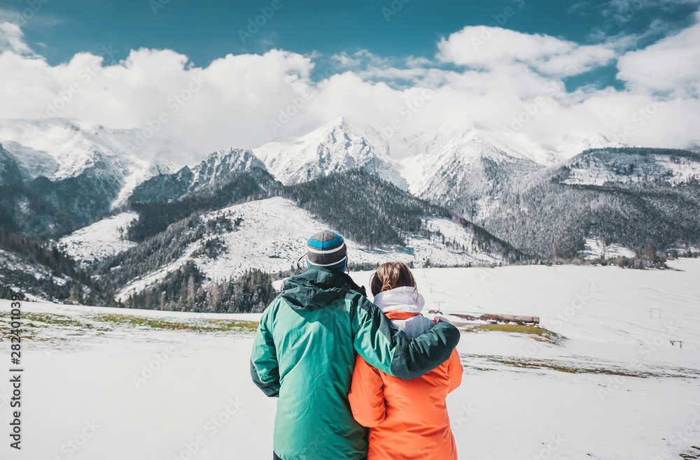 Back view photo of young loving couple hugging over winter mountains. Looking at mountains.