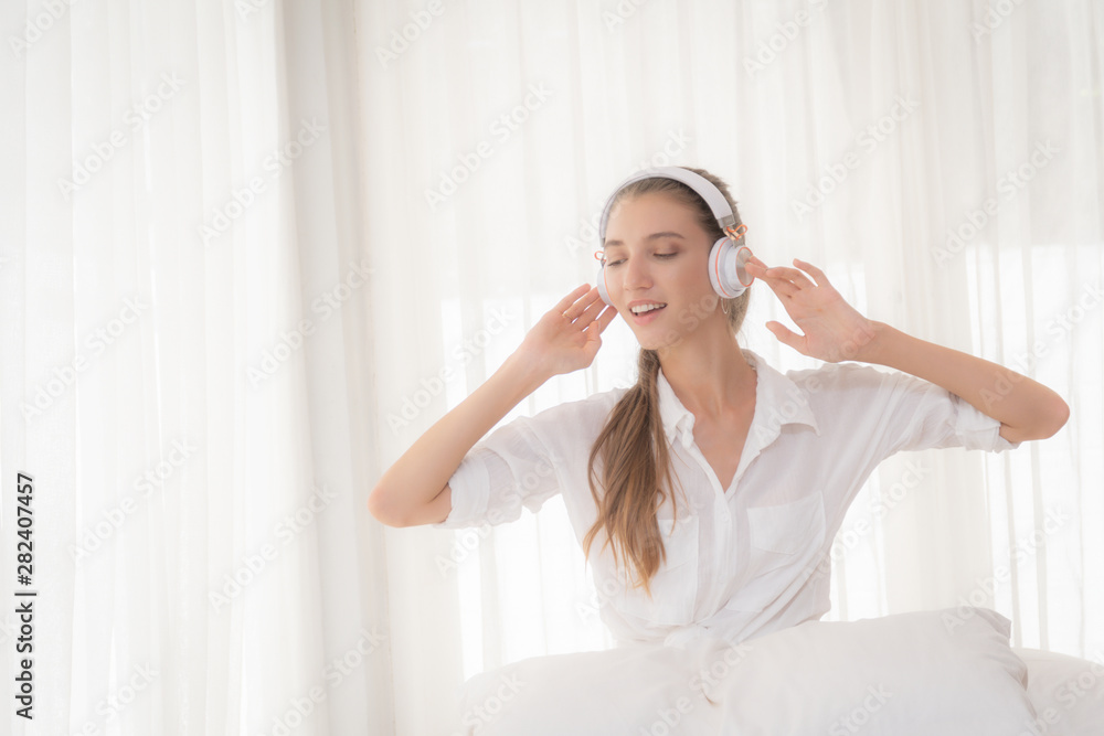 Beautiful woman long haired blond wearing a white shirt relaxing listening to music with headphones in the living room on white curtain background. Beautiful woman long haired blond wearing a white shirt relaxing listening to music with headphones in the living room on white curtain background.