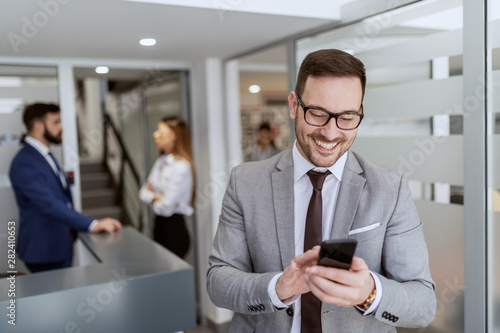 Young handsome smiling Caucasian businessman in suit talking using smart phone while standing in hall. In background his colleagues standing and chatting.