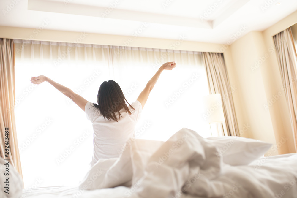 Woman stretching in bed after waking up, back view. Woman sitting near the big white window while stretching on bed after waking up with sunrise at morning, back view.