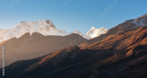 Beautiful mountains with snowy peaks at sunny morning in Nepal. Silhouettes of Himalayan mountains at sunrise. Colorful landscape with high rocks, blue sky and gold sunbeam. Amazing Himalayas. Nature