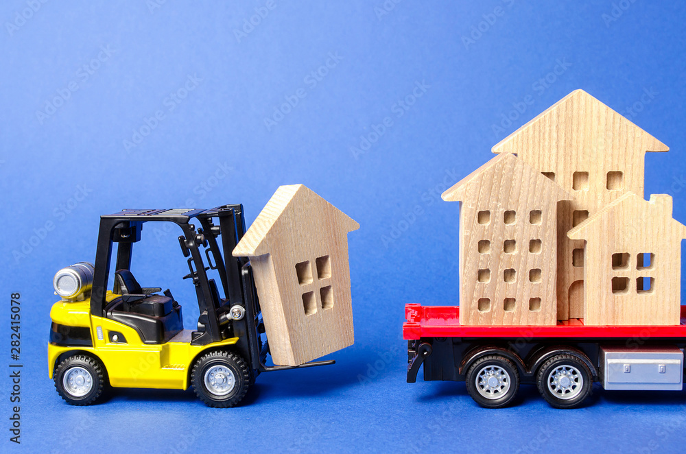 A yellow forklift loads a wooden figure of a house into a truck. Concept of transportation and cargo shipping, moving company. Construction of new houses and objects. Industry. Move entire buildings.