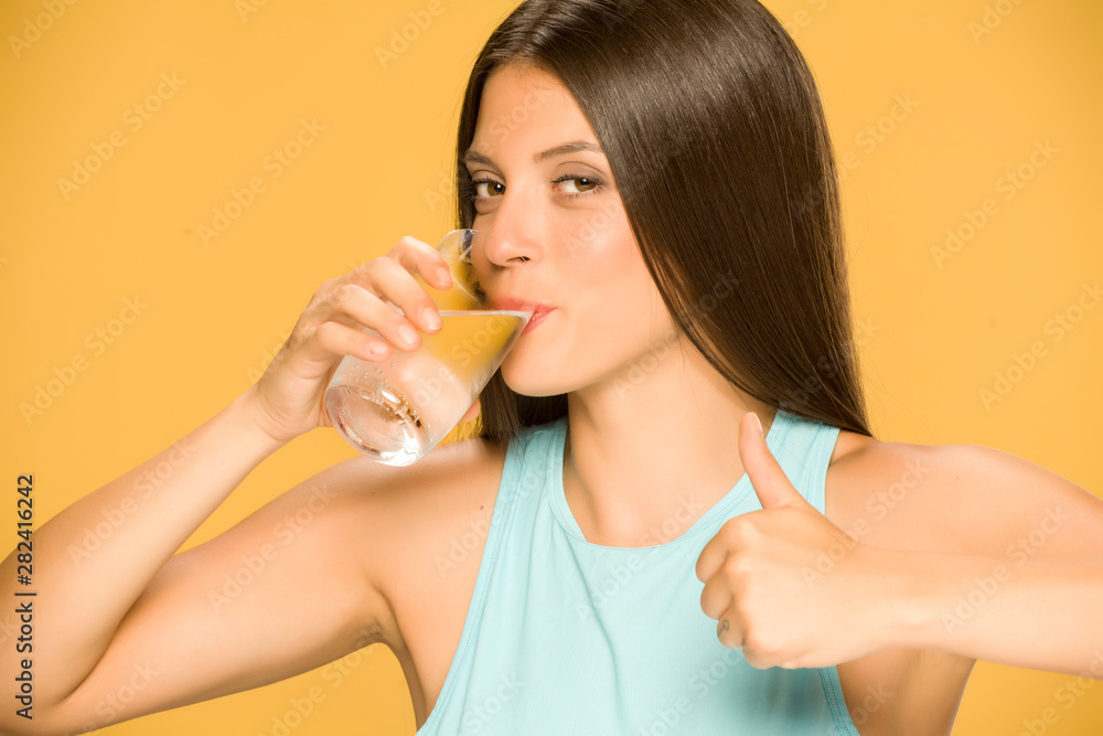 Young smiling woman drinking water from a glass and showing thumbs up on yellow background Young smiling woman drinking water from a glass and showing thumbs up on yellow background