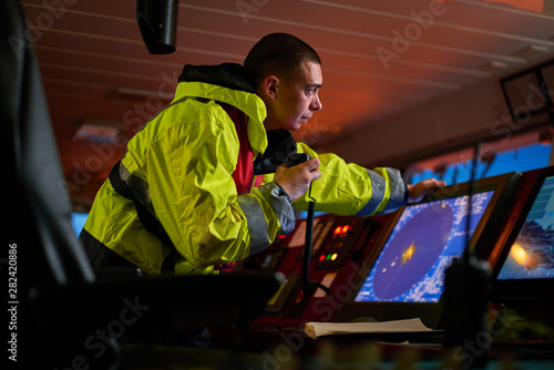 Navigator. pilot, captain as pat of ship crew performing daily duties with VHF radio, binoculars on board of modern ship with high quality navigation equipment on the bridge on sunrise.