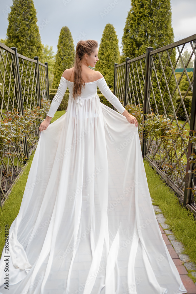 Fototapeta premium Princess in a vintage white dress. Walk along the picturesque summer field at sunset. A long train of white skirt fluttered on the run.