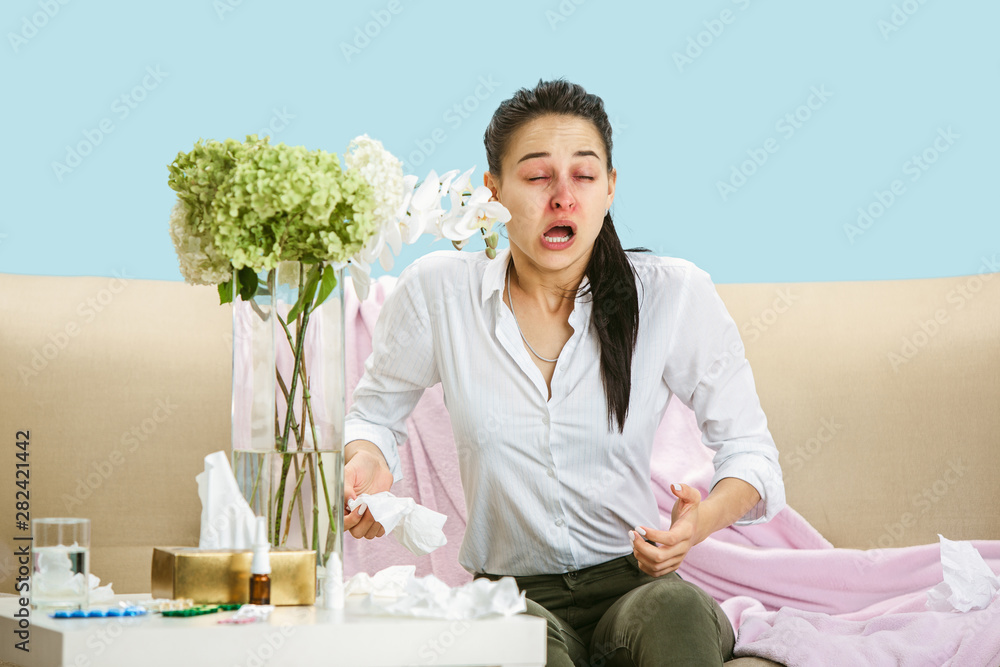 Young woman suffering from hausehold dust or seasonal allergy. Sneezing in the napkin and sitting surrounded by used napkins on the floor and sofa. Taking medicines with no result. Healthcare concept. Young woman suffering from hausehold dust or seasonal allergy. Sneezing in the napkin and sitting surrounded by used napkins on the floor and sofa. Taking medicines with no result. Healthcare concept.