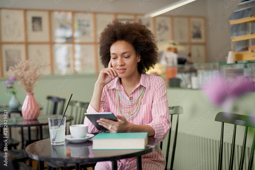 Attractive mixed race woman in striped pink dress sitting in cafe and using tablet.