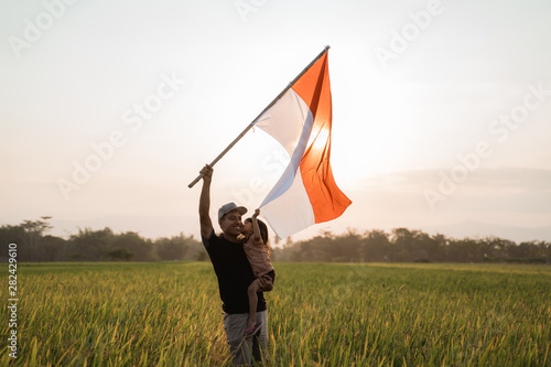 father carrying little girl pride flapping Indonesian flag with happiness in the rice field photo