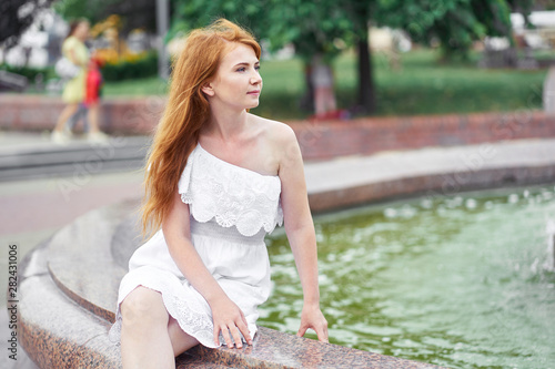 Attractive redhead girl sitting near the fountain in the city. Summertime.