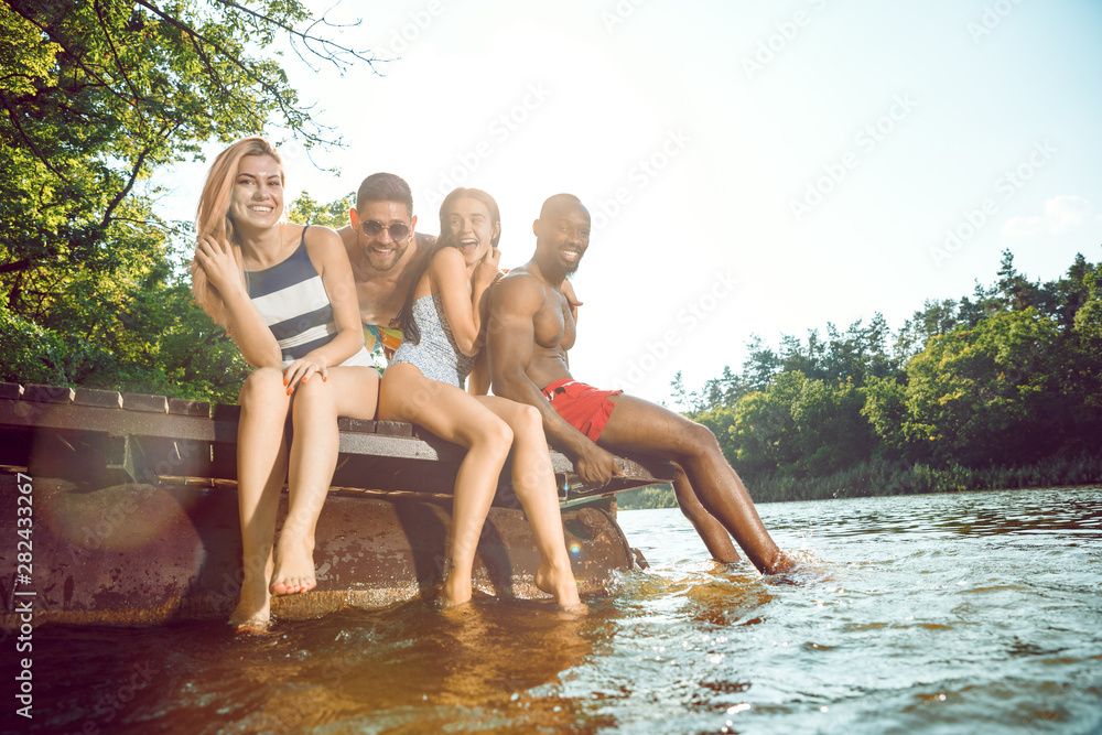 Fototapeta premium Group of happy friends having fun while sitting and laughting on the pier on river. Joyful women and men in swimsuit at riverside in sunny day. Summertime, friendship, resort, relations concept.