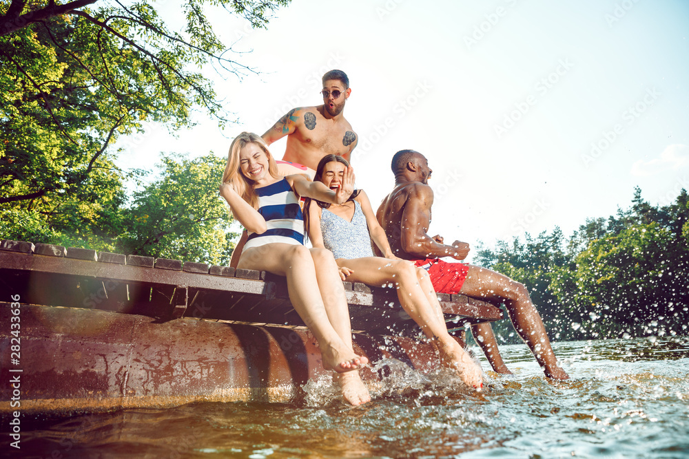 Fototapeta premium Group of happy friends having fun while sitting and laughting on the pier on river. Joyful women and men in swimsuit at riverside in sunny day. Summertime, friendship, resort, relations concept.