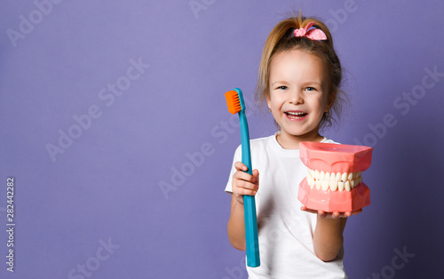 Obraz na płótnie Laughing kid girl shows us big dental implant model and giant toothbrush on purple with free text copy space w salonie