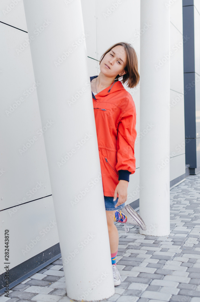 A short haired woman in coral windbreaker and colorful socks hiding behind a column near grey wall A short haired woman in coral windbreaker and colorful socks hiding behind a column near grey wall