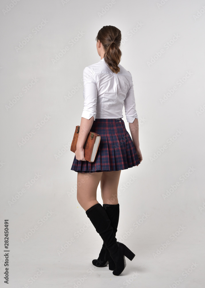full length portrait of a brunette girl wearing a white shirt and plaid skirt, standing pose and holding a book against a cream studio background. full length portrait of a brunette girl wearing a white shirt and plaid skirt, standing pose and holding a book against a cream studio background.