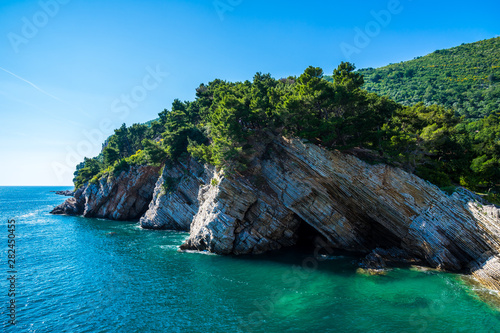 Montenegro  Rocky cliffs of petrovac bay coast covered by green trees and forest alongside azure water under blue sky