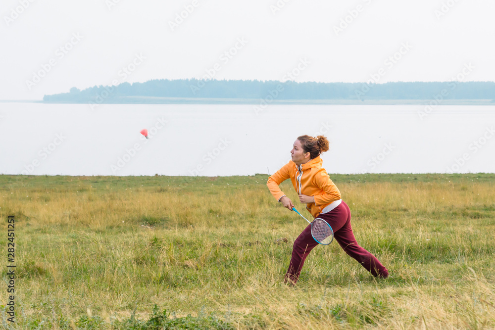 Beautiful girl in bright sportswear plays badminton on coast of lake.