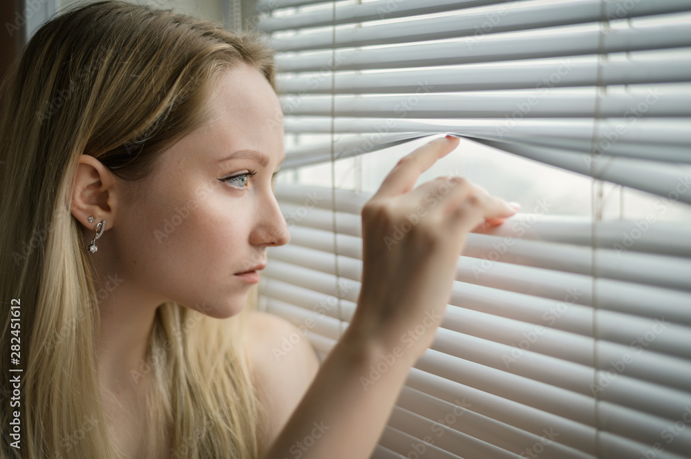 Young woman separating slats of blinds and looking through window.