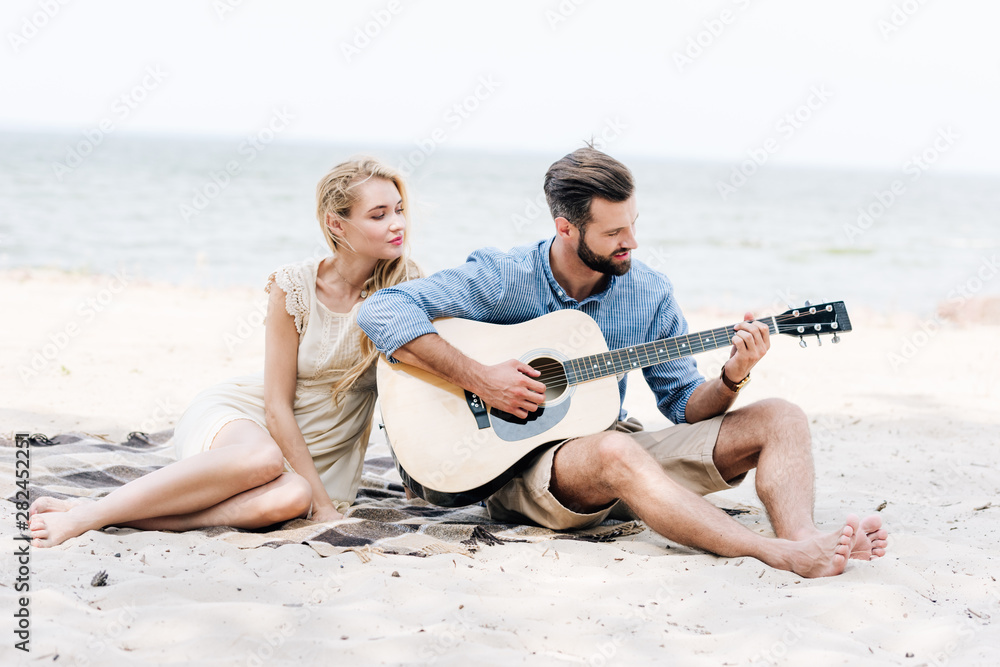 beautiful young barefoot woman sitting on blanket with boyfriend playing acoustic guitar at beach near sea