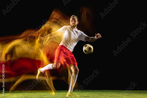 In fire. Young caucasian male football or soccer player in sportwear and boots kicking ball for the goal in mixed light on dark background. Concept of healthy lifestyle, professional sport, hobby.