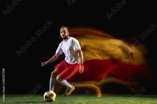 Team. Young caucasian male football or soccer player in sportwear and boots kicking ball for the goal in mixed light on dark background. Concept of healthy lifestyle, professional sport, hobby.