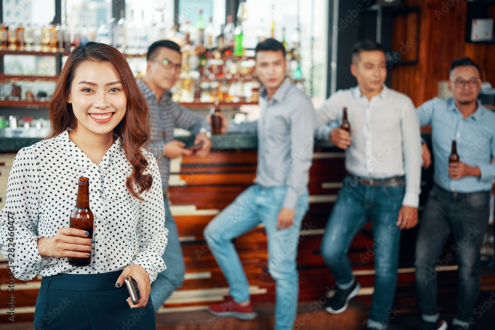 Obraz premium Portrait of Asian beautiful woman holding bottle of beer and smiling at camera with young men drinking beer at bar counter in the background