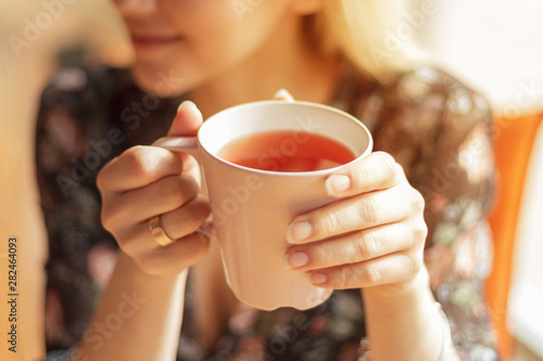 Close up photo of pretty woman holding mug of delicious hot tea, drinking, smiling, in cozy morning cafe, selective focus, noise effect