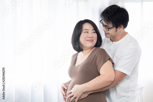 Lovely Asian adult pregnant couple happy hugging together with hand of husband touching belly of his wife