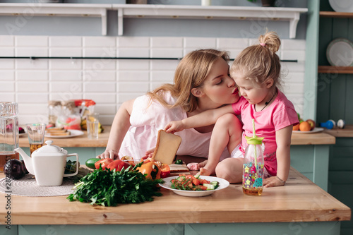 Side view of cute mother and daughter hugging and kissing each other in morning. Family sitting at kitchen, cooking breakfast and cutting vegetables at home. Concept of lunch and food.