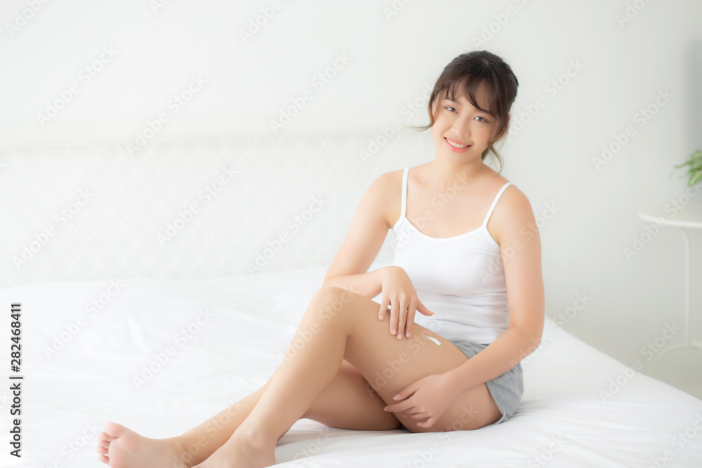 Beautiful young asian woman sitting on a bed stroking legs with soft smooth skin in the bedroom, girl applying touch body cream and lotion with treatment care, healthy and wellness concept. Beautiful young asian woman sitting on a bed stroking legs with soft smooth skin in the bedroom, girl applying touch body cream and lotion with treatment care, healthy and wellness concept.