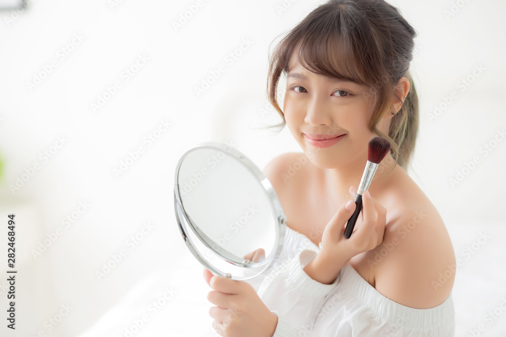 Beauty portrait young asian woman smile with face looking mirror applying makeup with brush cheek in the bedroom, beautiful of girl holding blusher, skin care and cosmetic concept. Beauty portrait young asian woman smile with face looking mirror applying makeup with brush cheek in the bedroom, beautiful of girl holding blusher, skin care and cosmetic concept.